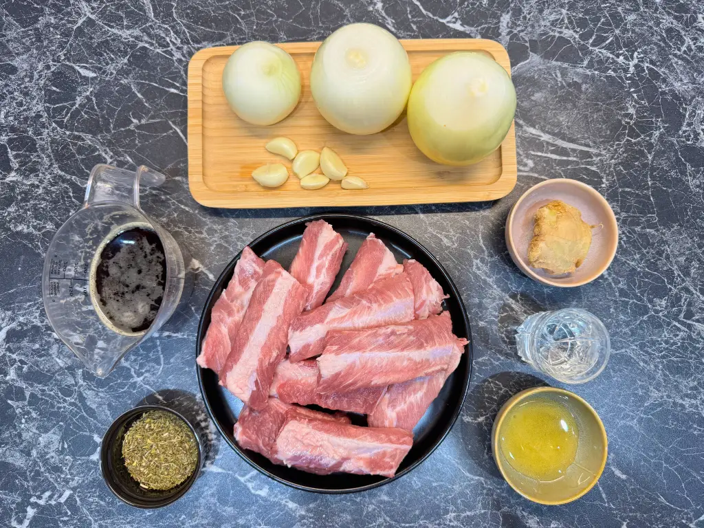 Raw pork ribs, onions, garlic, ginger, dark beer, honey, thyme, and water arranged on a kitchen counter before cooking.
