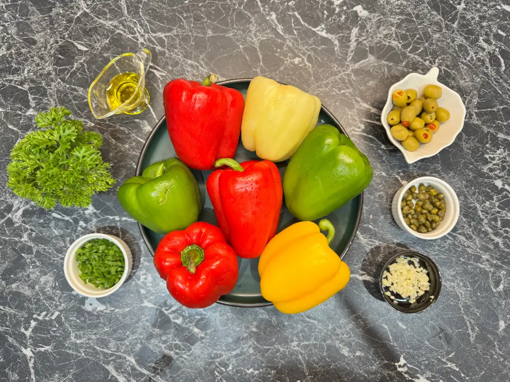 Fresh ingredients for roasted bell pepper and olive salad including peppers, parsley, garlic, olives, capers, green onions, and olive oil.