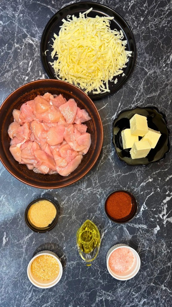 Flat lay of grated cheese, raw chicken pieces, butter, olive oil, smoked paprika, dried garlic, dried onion, and salt arranged on a dark marble surface.