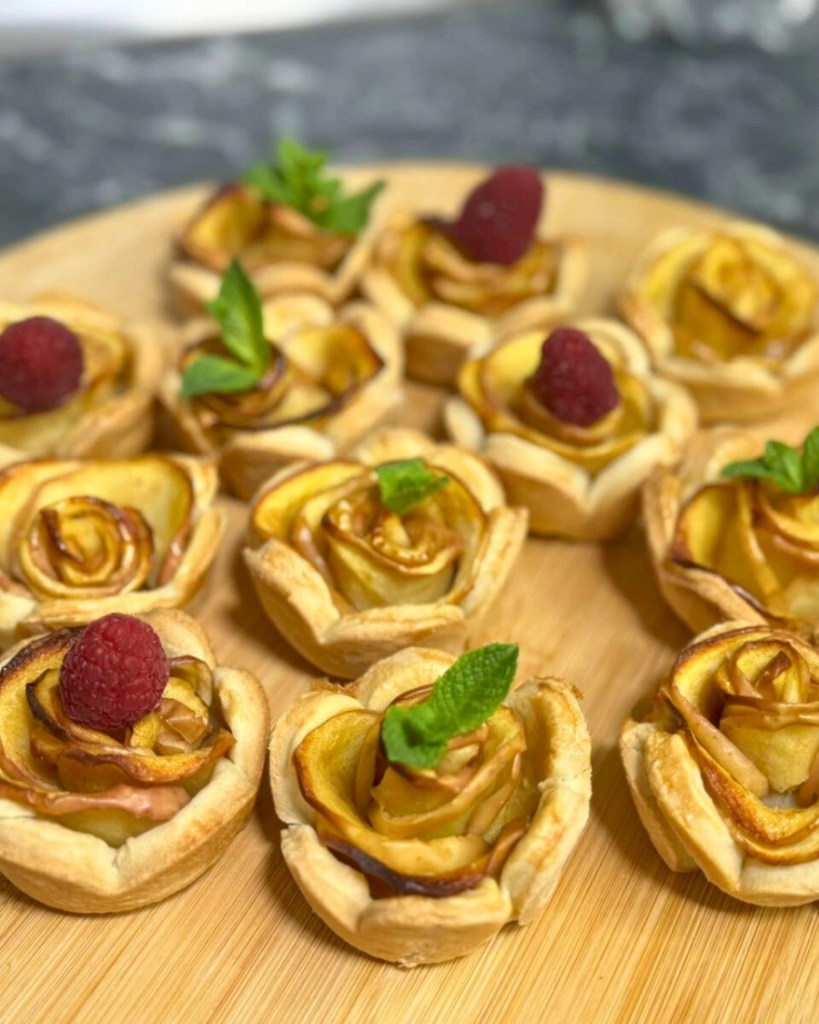 A tray of apple rose tartlets baked in heart-shaped puff pastry cups, decorated with raspberries and mint leaves.