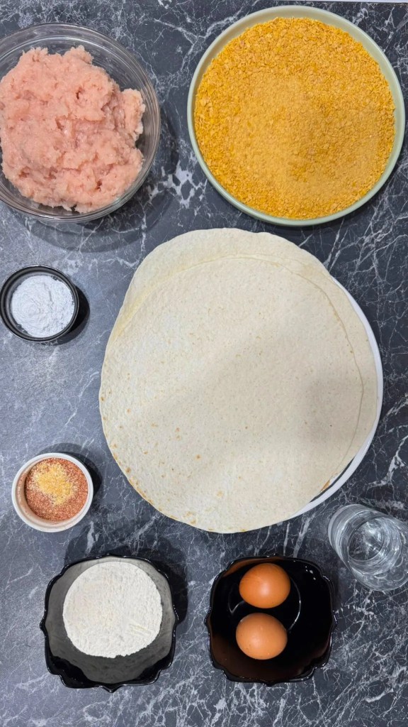 Flatlay of all ingredients needed to make Crispy Chicken Tortilla Bites, including chicken, tortillas, eggs, flour, and crushed cornflakes.