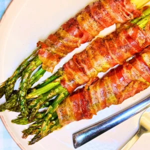 Oven baked bacon wrapped asparagus bundles served on a plate, with crispy bacon wrapped around fresh roasted asparagus spears.
