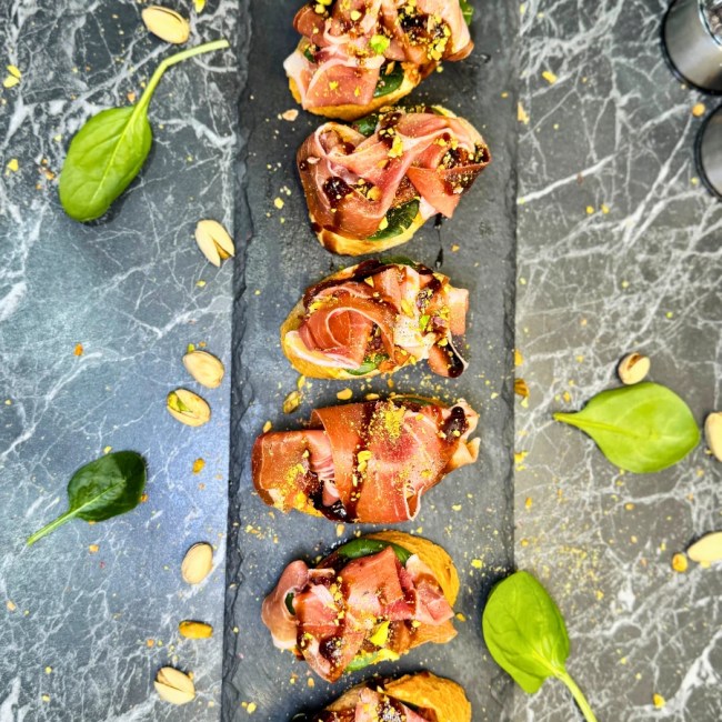 Overhead view of easy jamon serrano bruschetta topped with pistachios and balsamic glaze