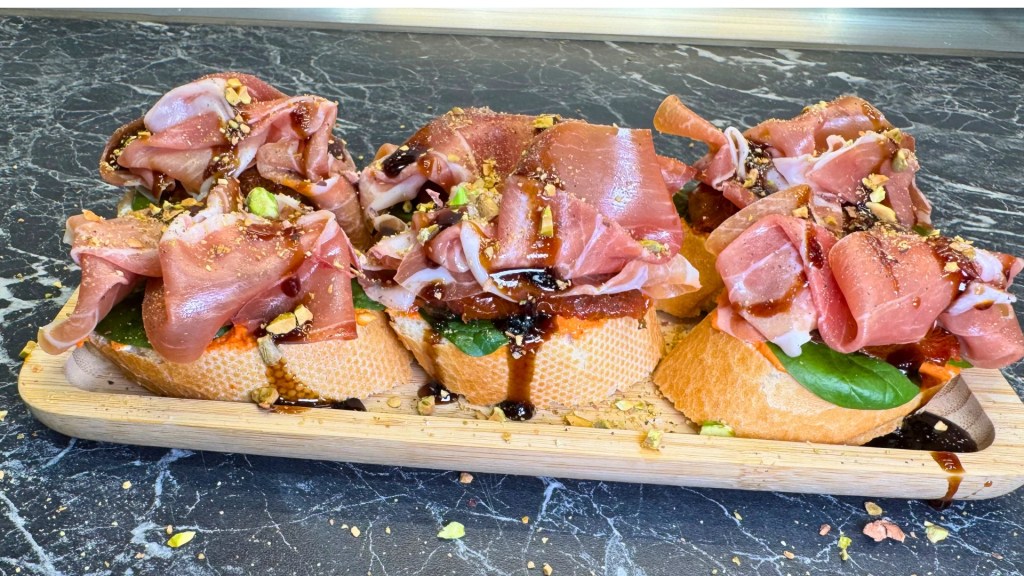Easy jamon serrano bruschetta topped with pesto, spinach, sun-dried tomatoes, pistachios and balsamic glaze