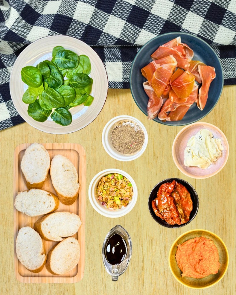 Ingredients for easy jamon serrano bruschetta including baguette, cream cheese, pesto, sun-dried tomatoes, spinach, pistachios and balsamic glaze