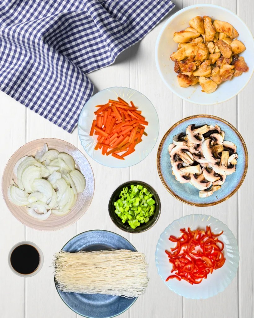 Flat lay of rice noodles, fried chicken pieces, sliced onion, carrots, mushrooms, red bell pepper, green onions, and soy sauce arranged on a white table.