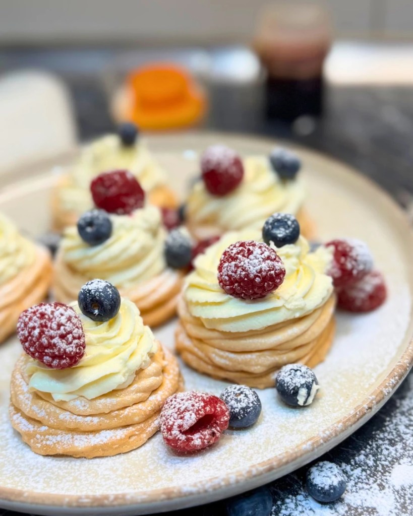 Mini pavlovas topped with mascarpone cream, raspberries, and blueberries, lightly dusted with powdered sugar on a serving plate.