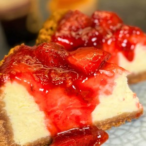 Slice of baked strawberry cheesecake with thick biscuit crust and fresh strawberry sauce dripping down the sides