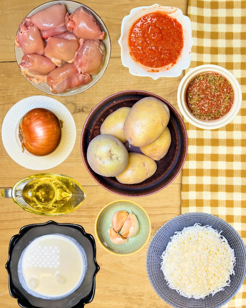 Ingredients for stuffed potatoes recipe including chicken thighs, potatoes, onion, garlic, tomato sauce, olive oil, spices, cream, and shredded cheese arranged on a wooden table.