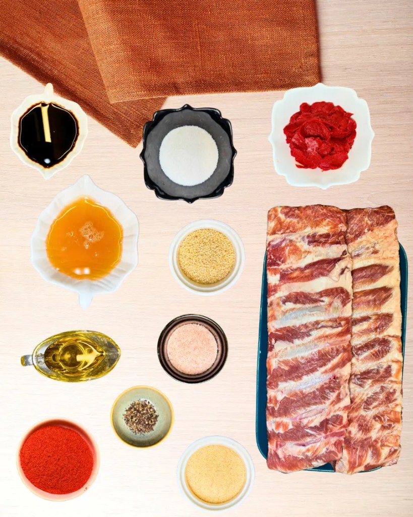 Ingredients for sticky honey soy pork ribs including pork ribs, soy sauce, tomato paste, honey, sesame seeds, olive oil, paprika, garlic powder, salt, sugar, and black pepper arranged on a table.