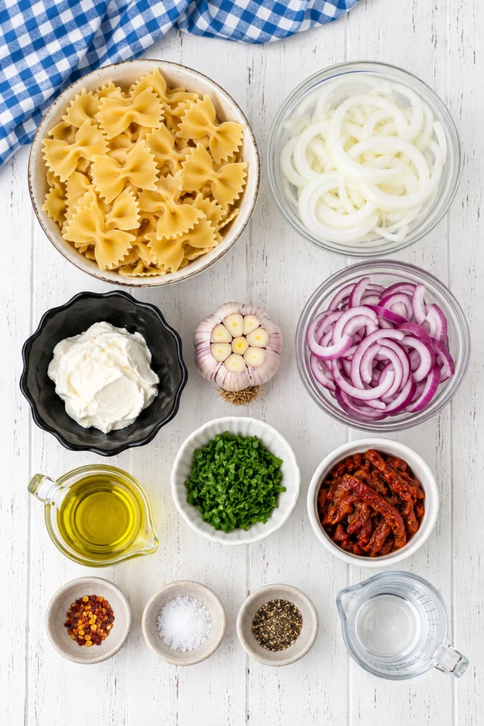 Ingredients for creamy onion pasta, including farfalle, sliced white and red onions, trimmed garlic bulb, cream cheese, sun-dried tomatoes, parsley, olive oil, and seasonings.