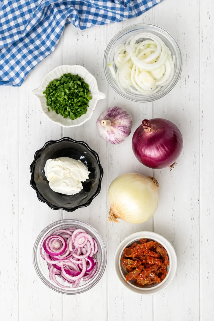 Ingredients for creamy onion pasta arranged on a white wooden table, including red and white onions, garlic, cream cheese, parsley, and sun-dried tomatoes.