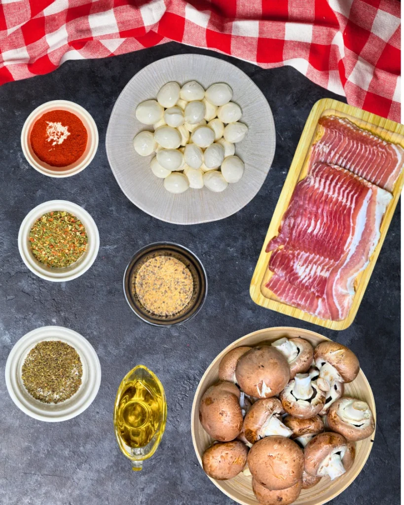 Fresh champignon mushrooms, baby mozzarella balls, sliced bacon, olive oil, garlic seasoning, dried herbs, and spices arranged on a kitchen countertop.
