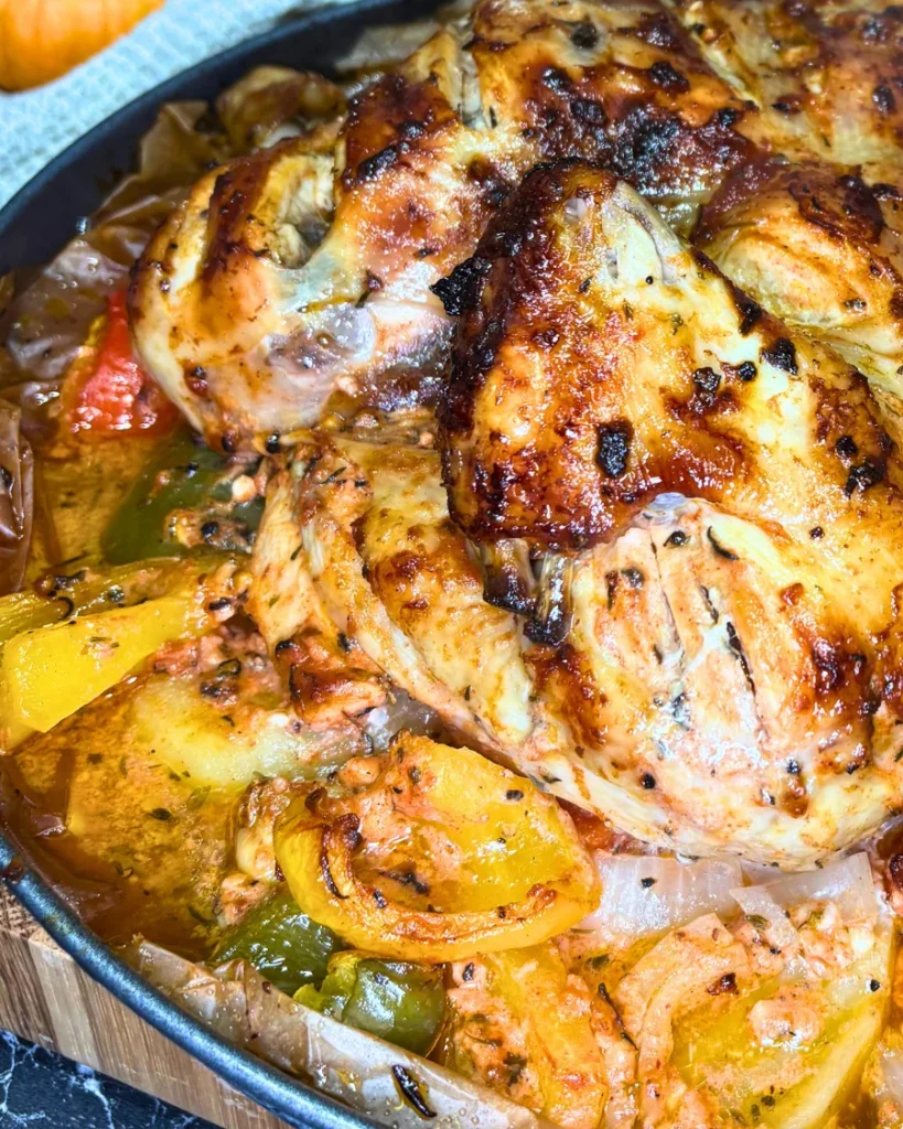 Close-up of golden-brown marinated roast chicken with tender potatoes, roasted garlic, and colorful bell peppers.
