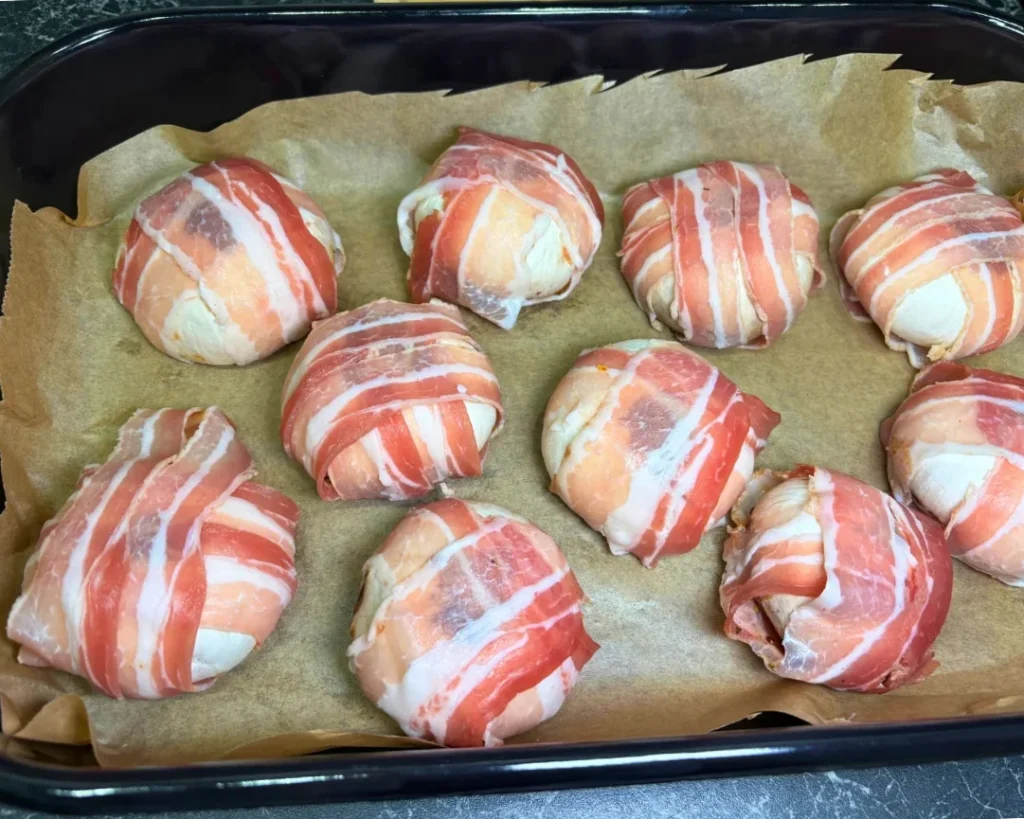 Raw mushrooms stuffed with mozzarella and wrapped in strips of bacon, arranged on a parchment-lined baking tray.